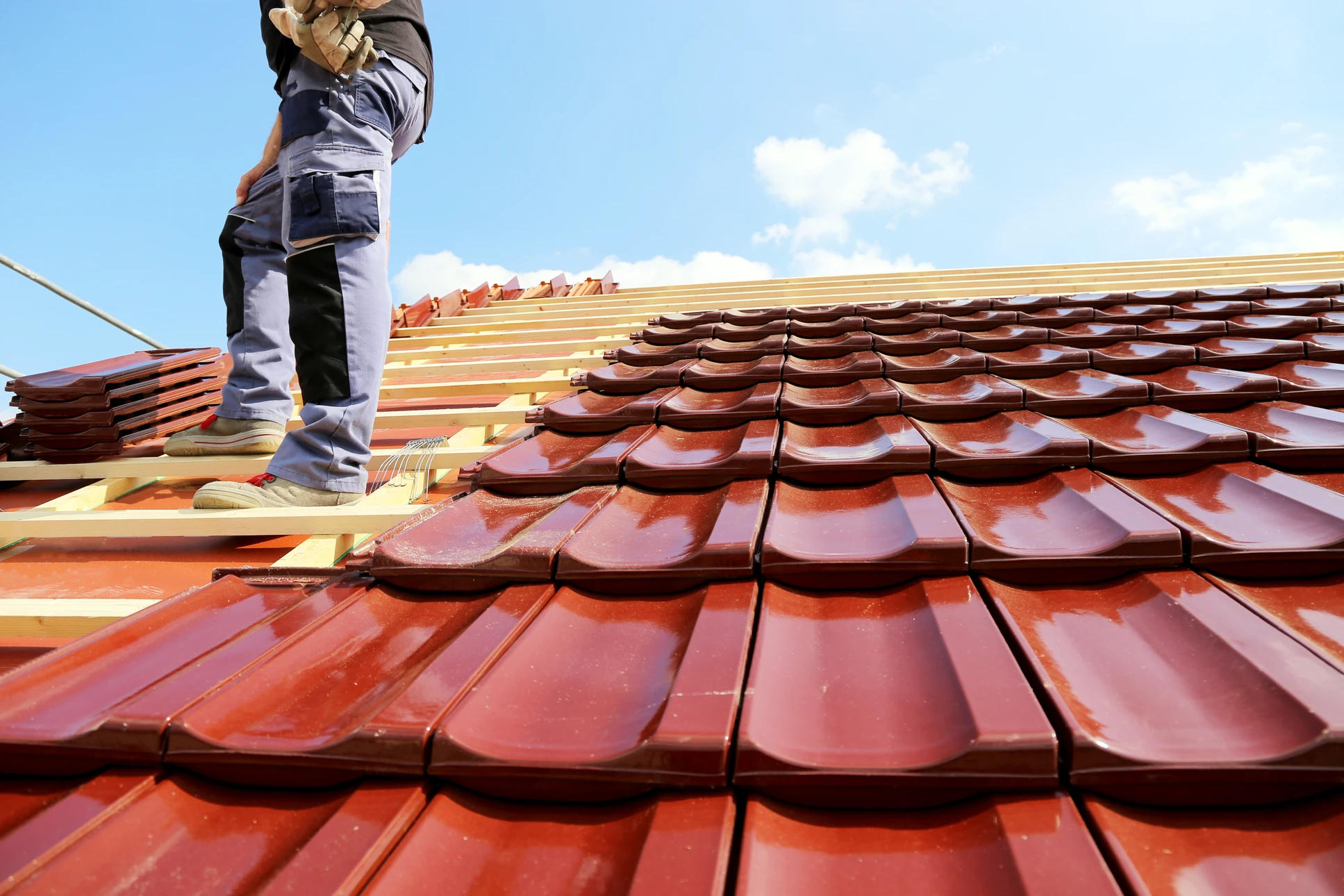 Ouvrier couvreur installant des tuiles terre cuite rouge sur charpente bois, travaux de toiture en cours sous ciel bleu