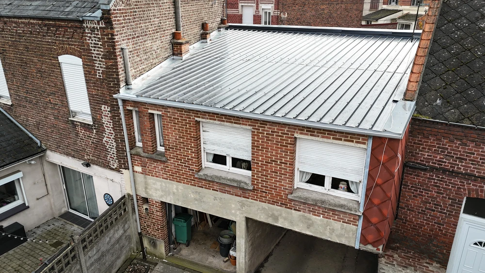 Toiture en bac acier nervuré zinc/alu, vue en plongée latérale, accolée à un mur pignon en briques rouges