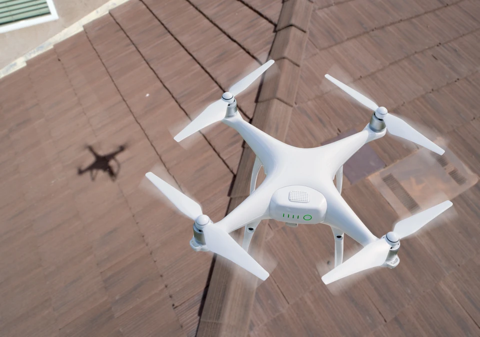 Photographie en extérieur montrant un drone blanc équipé de caméra professionnelle survolant une toiture résidentielle. Le drone est capturé en vol au-dessus du faîtage et des zones critiques de la couverture (tuiles, gouttières, noues), avec une maison visible en arrière-plan. L'image doit souligner la précision technique et la capacité d'inspection sans intervention physique, dans une lumière naturelle claire mettant en évidence les détails de la couverture inspectée.