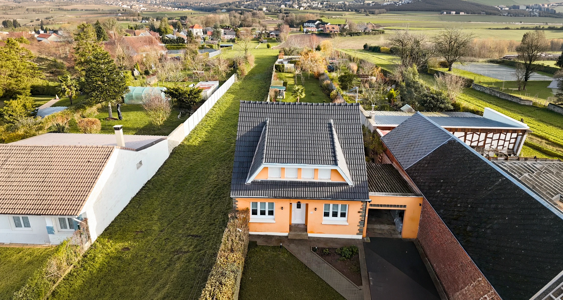 Un drone professionnel en vol au-dessus d'une maison résidentielle avec toiture en tuiles ou bac acier, capturant une vue aérienne rapprochée de la structure de toit et de la gouttière, dans une lumière naturelle claire de jour. L'image doit clairement montrer l'inspection aérienne d'une toiture depuis le ciel, illustrant la prestation de drone de vérification de toiture.